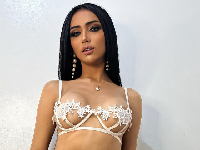 DannaWalton with long black hair, wearing a white lace bralette that reveals her small, perky tits and has floral embroidery. She's got pearl earrings and a heart necklace.
