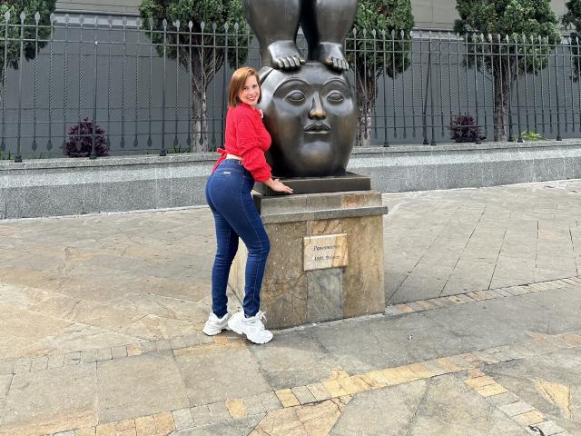 SaritaVelez with red shirt, tight blue jeans, white sneakers, standing by bronze statue of face with feet on top, hands on statue, curvy butt, short brown hair, smiling.