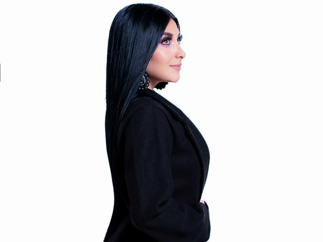 ArianaCortes with long black hair, wearing a black suit, side profile, looking forward, purple eye makeup, earrings, hands in pockets, white background, confident, sexy.