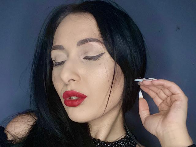 LexyClaire with black hair, red lipstick, and sharp winged eyeliner, eyes closed, hand with long white nails touching her hair, wearing a black beaded top, looking sexy.
