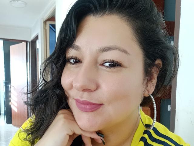 SakotoDika with dark wavy hair, pink lipstick, hoop earrings, and yellow striped shirt, resting chin on hand, smiling softly, brown eyes, light skin, casual indoor selfie.