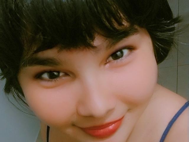 PrittyAlicia with short black hair, brown eyes, and red lipstick, smiling at the camera, wearing a blue spaghetti strap top, showing her cute face and slight cleavage.