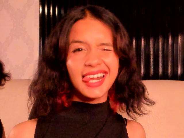 CatarsisNous, avec une peau marron moyen et des cheveux noirs ondulés, sourit avec la langue tirée, porte un haut noir sans manches et a des mèches rouges dans ses cheveux..