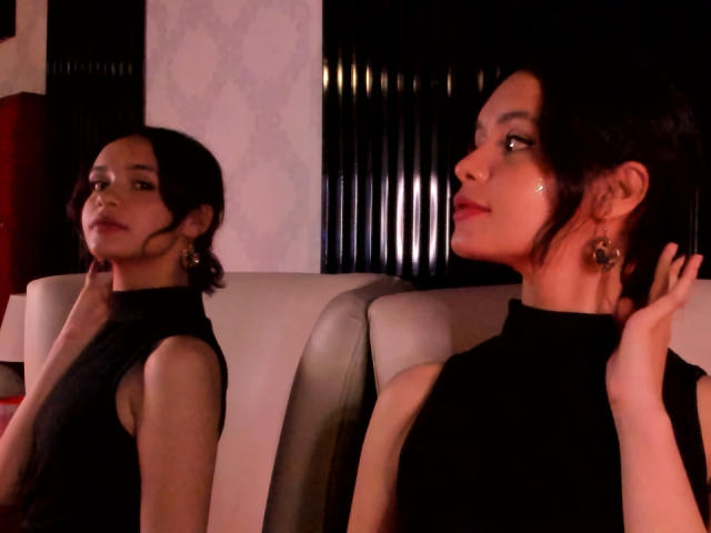 Dos mujeres CatarsisNous con cabello oscuro, vestidas con tops negros sin mangas, sentadas en una habitación con poca luz. Una CatarsisNous mira hacia adelante, la otra hacia un lado, con una mano levantada..