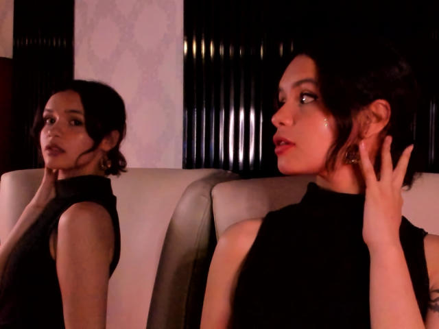 Due donne di CatarsisNous con capelli scuri, che indossano top neri senza maniche, sedute su un divano in pelle bianca, che guardano di lato, una con la mano vicino al viso, con una luce fioca e riflessi..