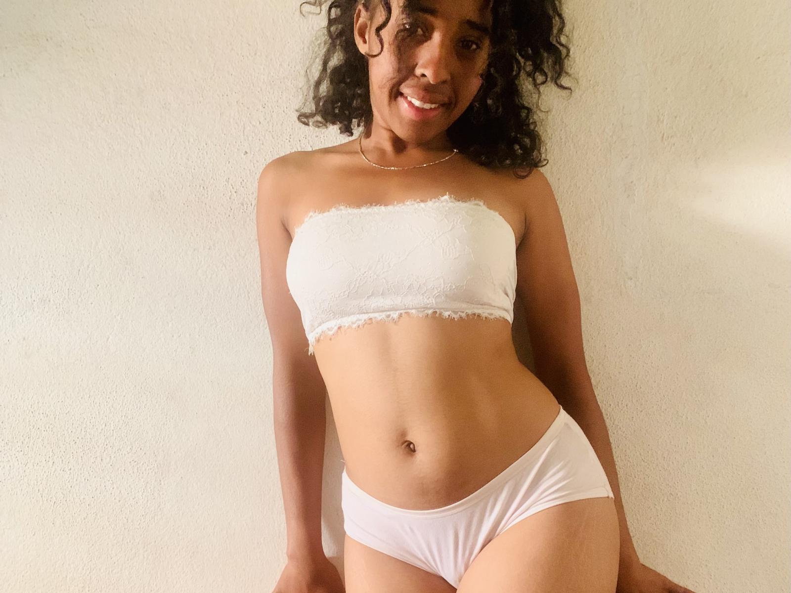 KittyHolms with curly hair, smiling, wearing a white strapless top and white panties, showing her slim, tan-skinned, flat-chested, small-boob, small-ass, flat-stomach, and belly button.