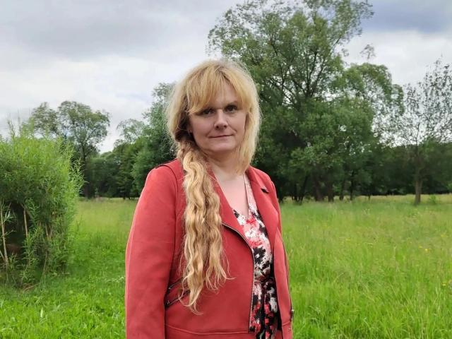 BeaLust-hot with long blonde hair, red jacket, floral dress, standing in green field, looking serious, grassy background, overcast sky, trees in distance, slightly wrinkled face, soft expression.