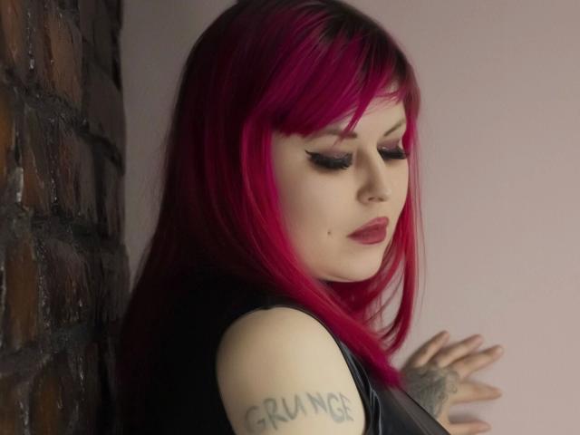 Psychobitch7: una ragazza con capelli rosa acceso, eyeliner scuro e rossetto rosso, appoggiata a un muro di mattoni, che mostra un braccio tatuato, guarda verso il basso e indossa una giacca di pelle nera..