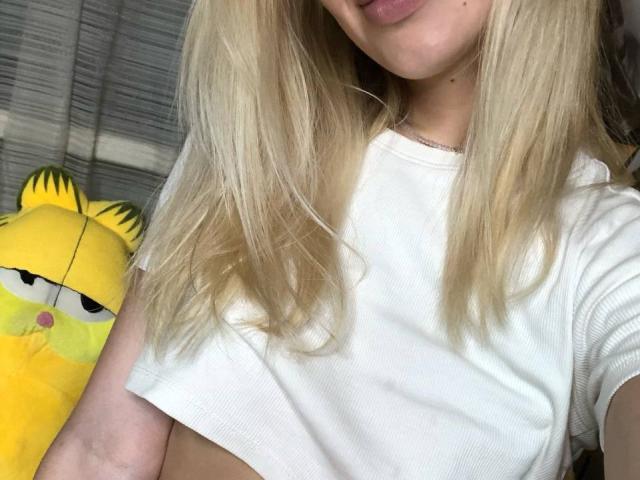 VanillaCake with blonde hair, wearing a white crop top, showing her tits, a yellow Garfield stuffed animal on the left, and a subtle smile.