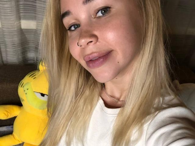 VanillaCake has blonde hair, a nose ring, and a subtle smile, wearing a white shirt, with a yellow stuffed Garfield on the left. Soft light, cute face.