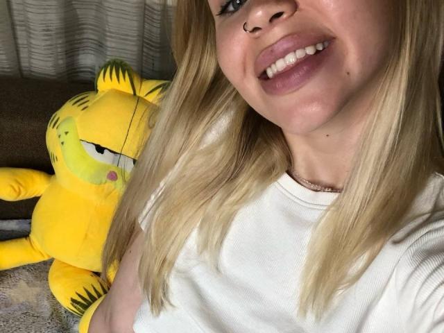 VanillaCake with blonde hair, white shirt, and pink lips, smiling at a yellow Garfield plushie on a brown couch. Her nose is pierced.