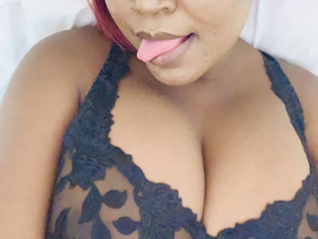Rynah's brown skin and pink tongue are out, wearing a black lace bra that shows off her big tits. She has red hair and a cute face.