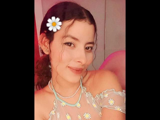 Elena Santini, avec des cheveux bruns, une fleur de marguerite dans les cheveux, un haut transparent à épaules dénudées avec des motifs de marguerites, un collier de perles, souriante, mignonne, poitrine de taille moyenne, éclairage rose doux..