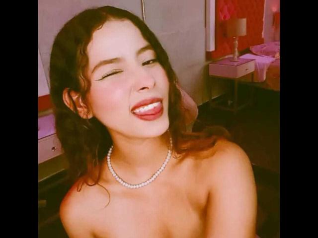Elena Santini, avec ses cheveux bruns bouclés, souriant avec la langue tirée, un collier de perles, les épaules dénudées, des lèvres roses douces et un maquillage léger, sur une photo légèrement floue aux tons chauds..