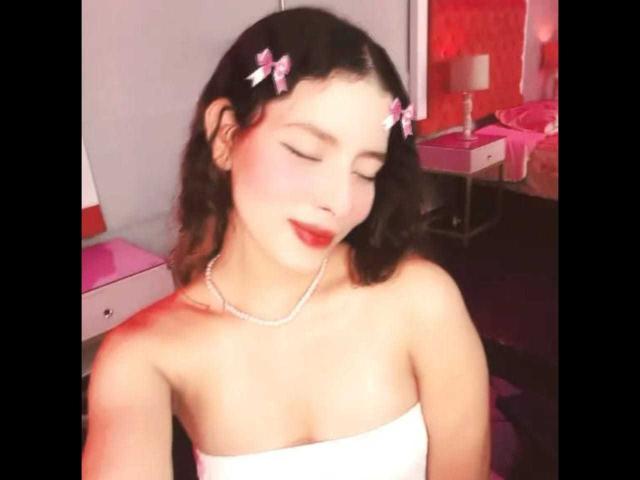 Elena Santini hat niedliche rosa Haarspangen, roten Lippenstift und ein weißes Top ohne Träger, das ihre kleinen Brüste zeigt, sowie eine Perlenkette. Ihre Augen sind geschlossen und sie hat ein sanftes Lächeln..