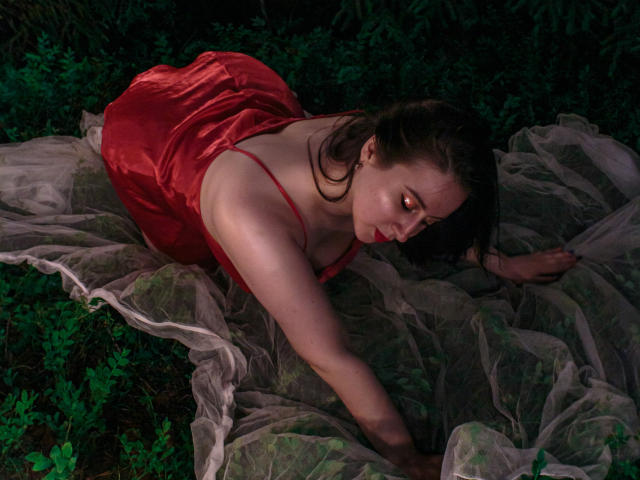 ViaLattea on her knees, wearing a red satin dress, with a sheer gray fabric beneath her. She has pale skin, dark brown hair, and a focused expression.