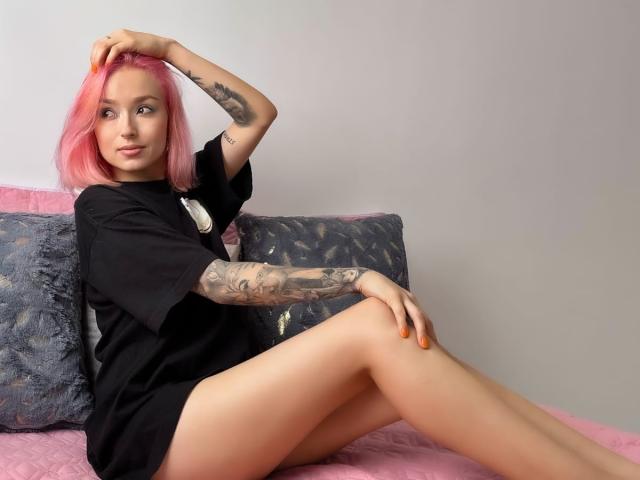 SarahRousen with pink hair, tattoos on arm, black t-shirt, sitting on pink bed, legs crossed, hand in hair, orange nails, looking at camera, smiling, cute, sexy.