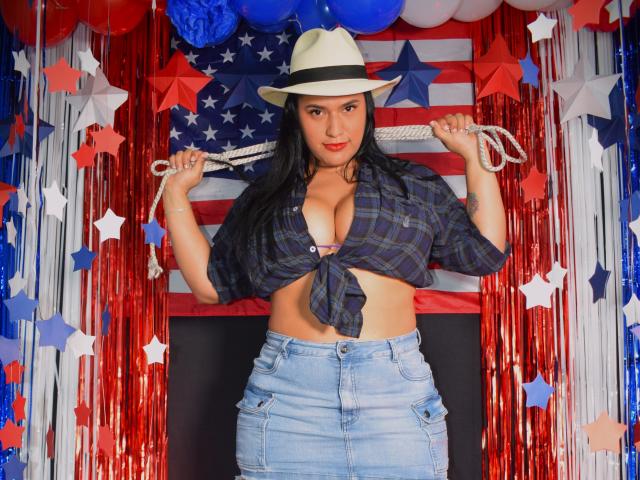 KorinaKova with big tits, wearing a tied-up plaid shirt, blue jeans, and a white hat, holding rope, standing in front of American flags and red star decorations.