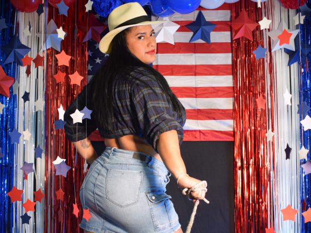 KorinaKova with long black hair, wearing a plaid shirt, denim shorts, and a beige hat, holding a rope, showing her ass, and looking over her shoulder.