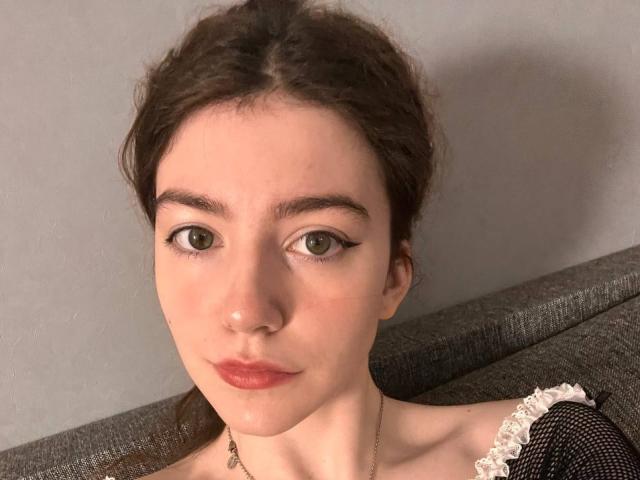 VinnaSkyHott with brown hair, green eyes, red lipstick, and a cute nose, looking straight at the camera. She's wearing a black lace top and a thin necklace.