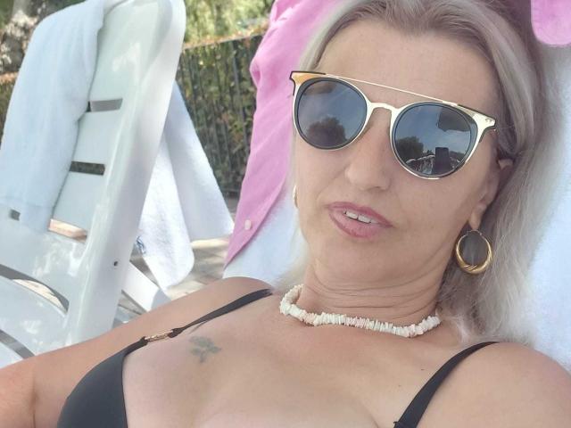 SexyCoco con cabello rubio, gafas de sol doradas, sostén negro, collar de conchas blancas, aretes de aro dorados, un tatuaje pequeño en el seno derecho, una toalla rosa detrás, una silla blanca y una sonrisa relajada..