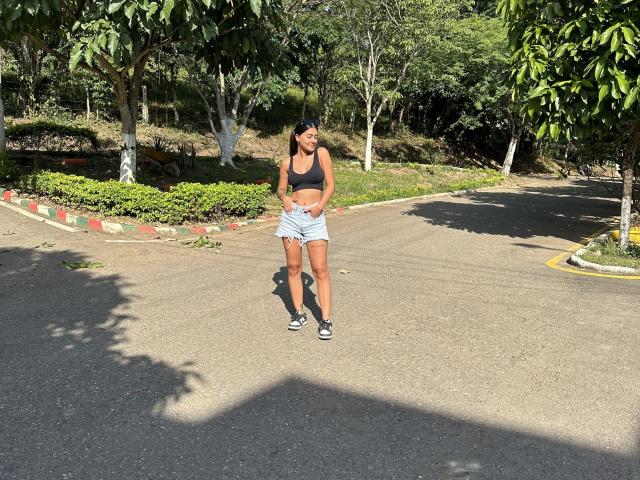 ThaliaAres with brown skin, black tank top, small boobs, white shorts, black socks, and sneakers, standing on a sunny road, hands on hips, looking down, trees in background.
