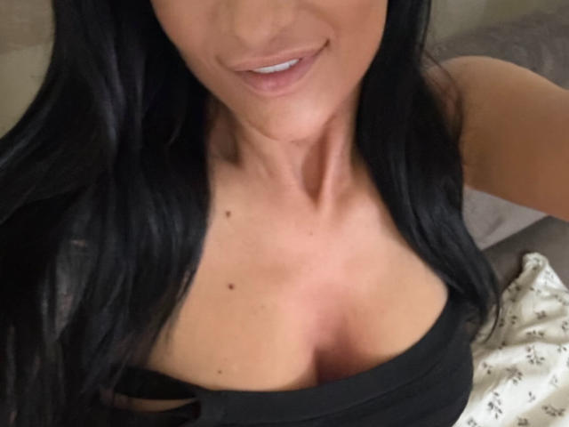 AndraD's upper body, showing black hair, slight smile, and cleavage in a black top with visible tits and small moles on her chest. She has light skin and blue lips.