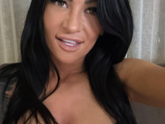 AndraD has long black hair, brown eyes, and is smiling with her teeth slightly showing. She's topless, with visible cleavage. Her skin is tan, and she's taking a selfie.