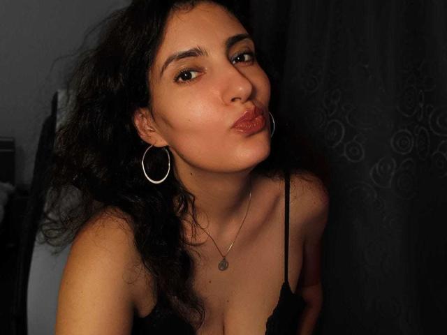 AddictedToUTS with curly black hair, hoop earrings, and a black lace top, pouting with slightly parted lips, showing subtle cleavage and a small necklace.