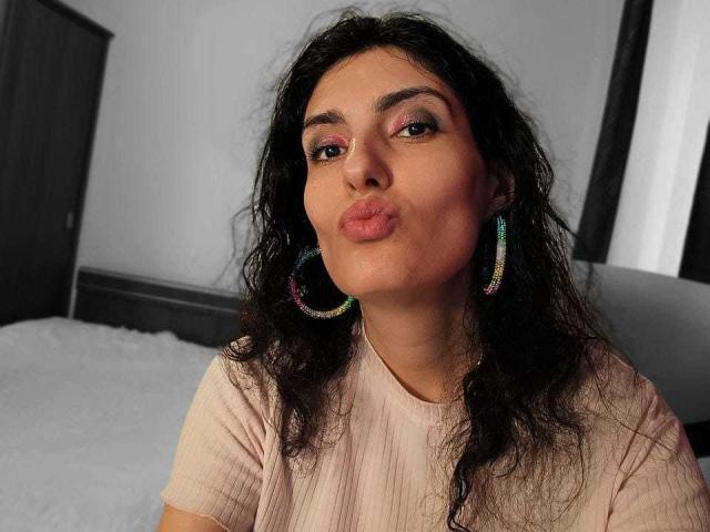 AddictedToUTS with curly black hair, pink shirt, colorful hoop earrings, and pouty lips, standing in a gray room. Her brown eyes are slightly open and she's looking forward.