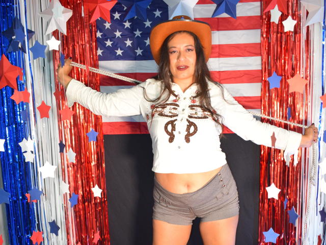 MiaCallie, con cabello largo y castaño, usando un sombrero de vaquera marrón, una camisa blanca con un diseño decorativo marrón y pantalones cortos marrones, sosteniendo una cuerda, mostrando su abdomen plano y sus glúteos pequeños..