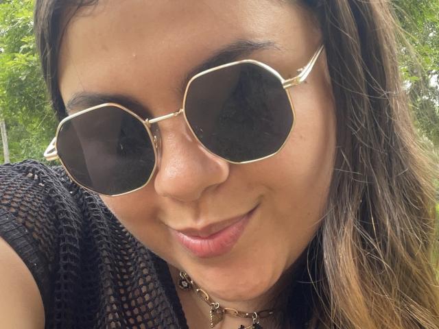 MissSofiia with brown eyes, wearing gold aviator sunglasses, black mesh top, and gold chain necklace, smiling with slightly parted pink lips. Her brown hair is loose.