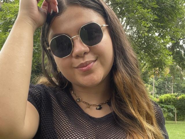 MissSofiia with round sunglasses, brown wavy hair, black mesh top, necklace, and a subtle smile, touching her head. Her tits are partially visible through the mesh.