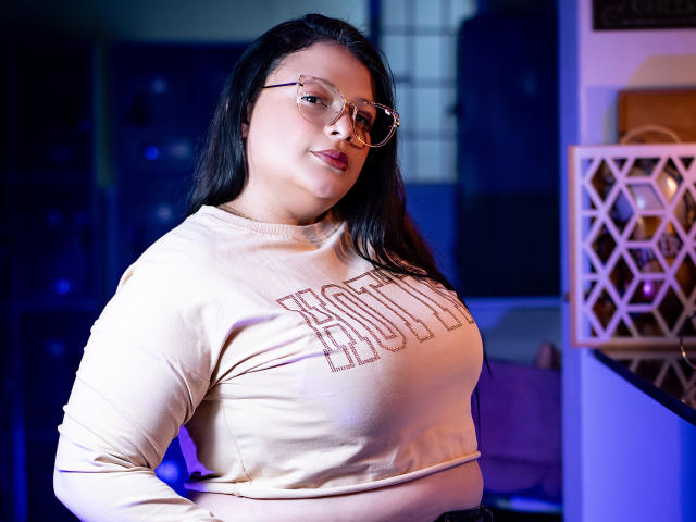 SherylHamilton with glasses, medium skin, black hair, large tits in a beige long-sleeve shirt, standing confidently, blue and purple lighting, slightly smiling, hands on hips.
