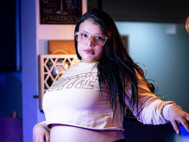 SherylHamilton with large tits, wearing a beige long-sleeve crop top, glasses, and long black hair, standing with arms out, blue and purple lighting in the background.