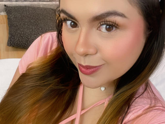MissSofiia has pink lipstick, long brown hair, brown eyes, and a small heart necklace. She's wearing a pink top and has a cute smile. Her tits are not visible.
