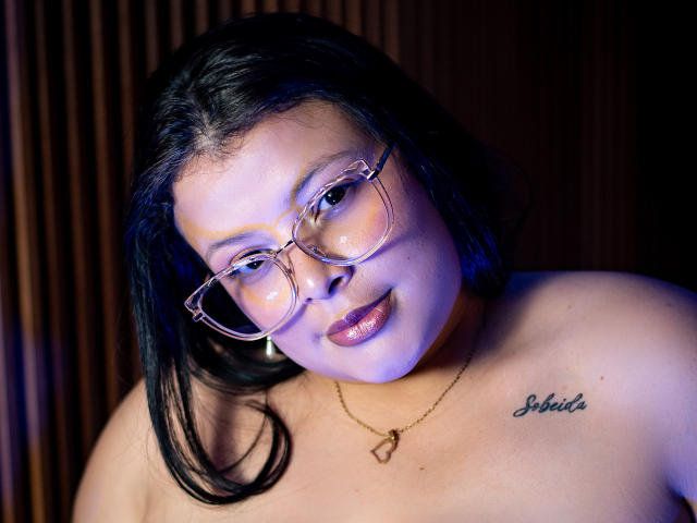 SherylHamilton has shiny black hair, glasses, and a tattoo reading "Sabrita" on her shoulder. She's topless, with her tits exposed, and a gold necklace. Blue lighting highlights her.