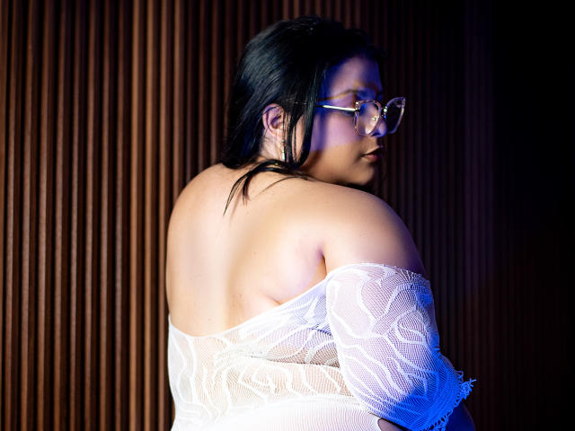 SherylHamilton with glasses, black hair, and white lace top, showing her back and bare shoulders, looking over her shoulder, highlighting her ass and smooth skin.