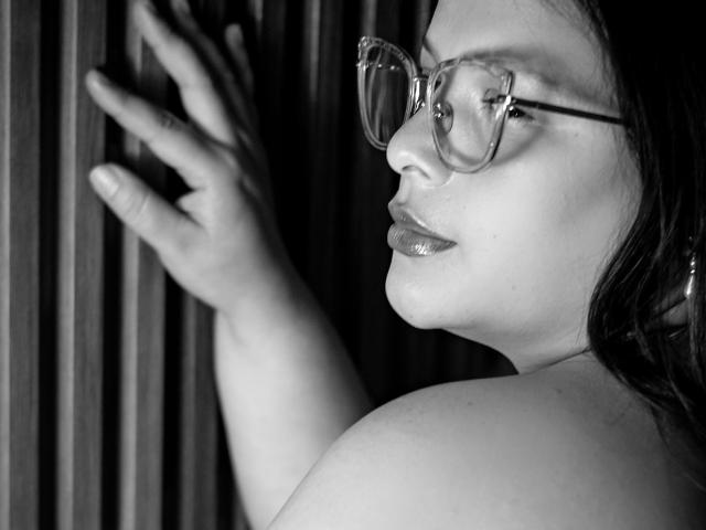 SherylHamilton with glasses, shiny lips, touching vertical wooden slats, her hand and shoulder visible, looking sideways, monochrome photo.