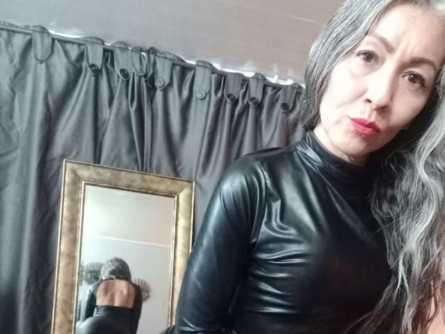 Nicolette Shea, com cabelos grisalhos longos, vestindo um cachecol de couro preto, com lábios vermelhos franzidos, e mostrando os seios em um reflexo no espelho. Seu traseiro está ligeiramente visível..
