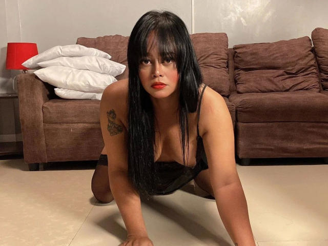 YENDYSFOX01-hot, black-haired, red-lipstick, tattooed, wearing black lingerie, on knees, hands on floor, looking at camera, brown couch, white pillows, red lamp in background.