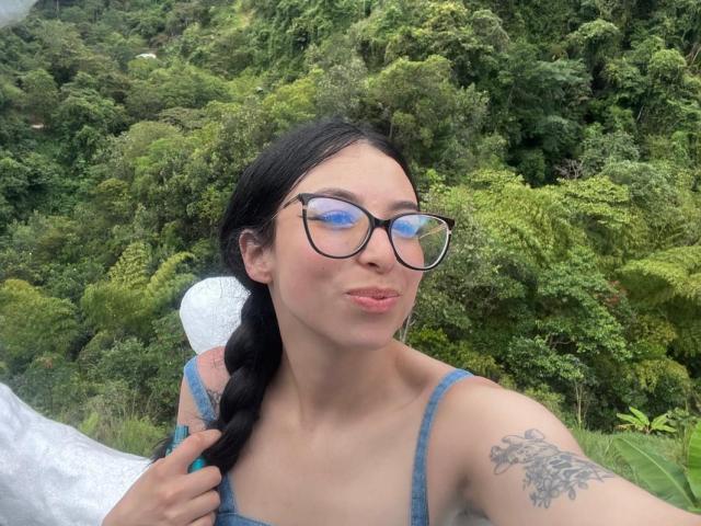 CamilleRios with glasses, pouty lips, black braid, blue top, tattoo on right arm, holding white bag, green forest background. Boobs, tits, ass, pussy implied by top.