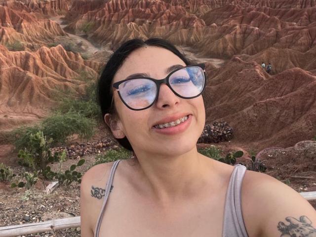 CamilleRios usa óculos pretos, tem um sorriso que mostra os dentes e uma tatuagem no ombro direito. Ela está vestindo uma camiseta cinza sem mangas e está em um deserto com formações rochosas vermelhas..