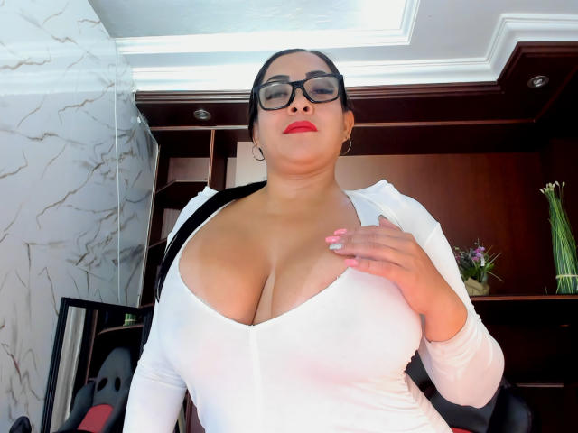 MellissaCortez with glasses, red lipstick, and white top, showing large tits, hand on chest, pink nails, black hair, in office, wooden shelves, vase of flowers in background.