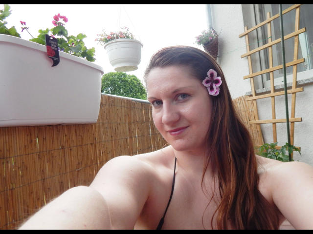 Sexyanny88-hot with long brown hair, pink flower hairpin, black halter top, smiling, taking selfie, showing her boobs, in bamboo-tiled outdoor space with hanging planters.