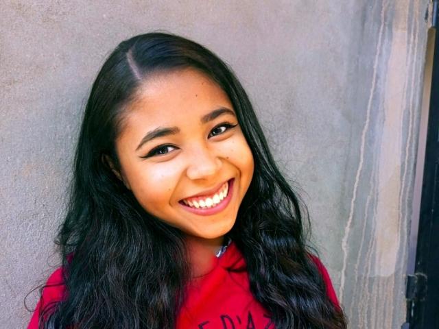 Mayumii with long black wavy hair, smiling widely, showing white teeth, wearing a red shirt, standing against a gray wall, has dark eyes and subtle makeup.