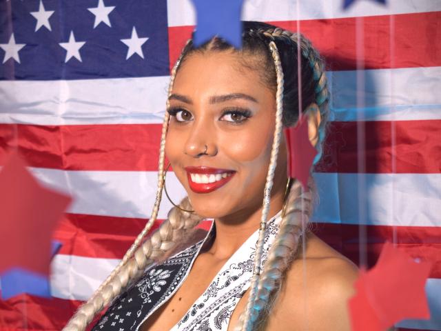 LilithWalker with blonde braids, red lipstick, hoop earrings, black and white halter top, smiling at camera, American flag in background, shiny skin, cute, sexy, bright lighting.