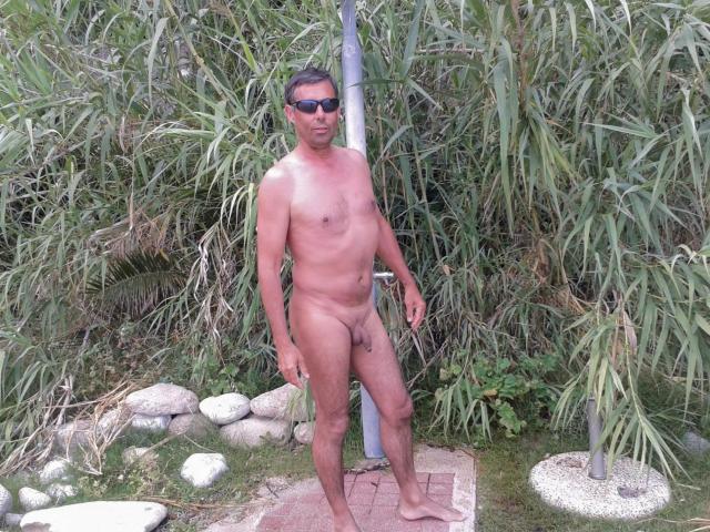ActifMan, nu, se tient debout avec un pénis flasque, portant des lunettes de soleil sombres, devant de hautes herbes vertes, avec des rochers et un chemin de gravier. Il a un corps de taille moyenne, poilu et bronzé..