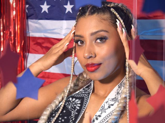 LilithWalker hat blonde Zöpfe, roten Lippenstift und berührt ihre Ohren mit erhobenen Händen. Sie trägt ein schwarz-weiß gemustertes Oberteil, im Hintergrund ist eine amerikanische Flagge zu sehen..