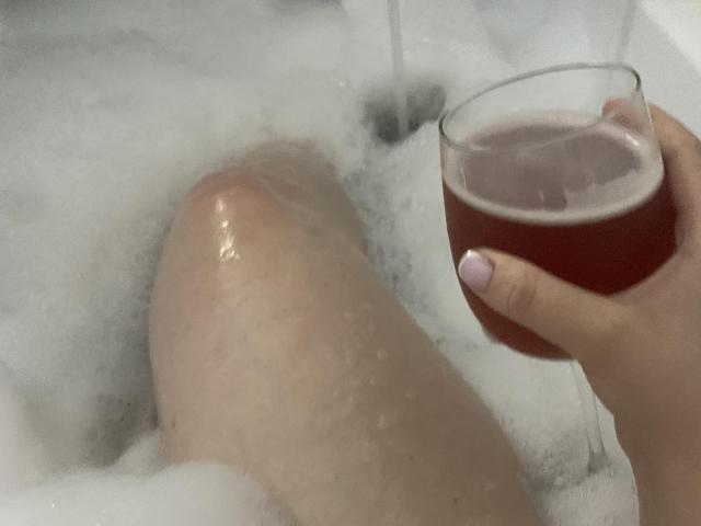SharonStorm's wet knee in a bubble bath, holding a glass of pink drink with a pink-nail-ed hand.